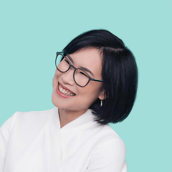Headshot of Prae Songprasit facing left and smiling widely. She has short shoulder length black hair, and is wearing glasses with a white top. The image sits on a plain Aqua Spray background.